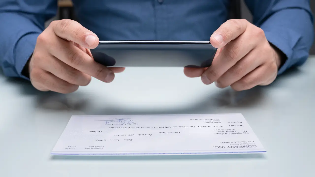 A person holding a paper check while using a smartphone banking app to attempt a mobile deposit at a desk