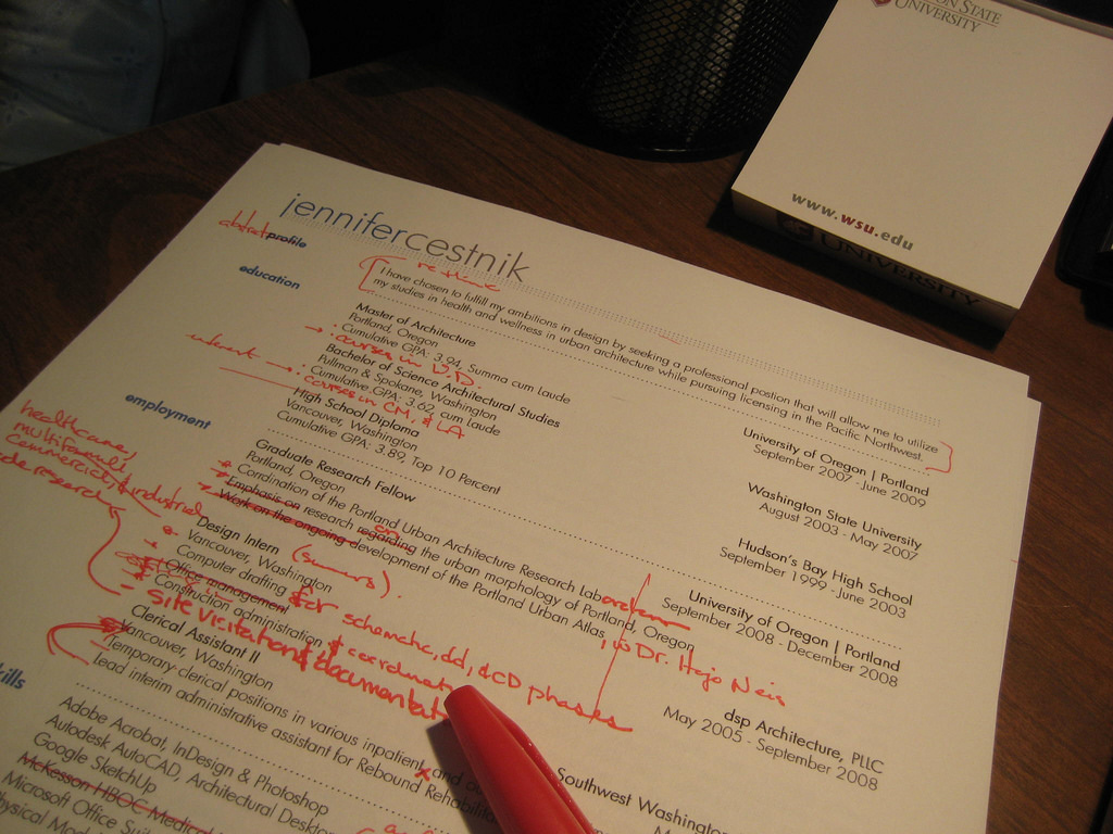 A person reviewing printed resume pages with handwritten notes on a desk beside a laptop.